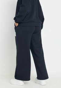 Person med löst sittande svarta joggingsbyxor och en matchande sweatshirt, står med en hand i fickan, synliga vita sneakers på vit golv.