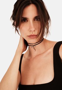 Sort choker-halskæde med to tynde snore og en sølvkors vedhæng, båret mod en ensfarvet baggrund, der fremhæver halsudskæringen og kravebenet.