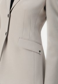 Béžový přizpůsobený blazer se strukturovaným střihem, s vertikálními švy, jednou náprsní kapsou a detaily loga na klopě kapsy.