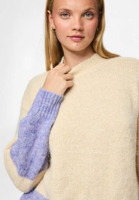 Pull en maille avec un corps beige et des manches lavande. Tissu texturé, encolure ronde et poignets côtelés. Apparence douce et chaleureuse.