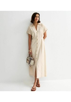 Kobieta w beżowej, midi sukience na guziki z krótkimi rękawami, trzymająca małą torebkę z przywiązaną apaszką, nosząca brązowe sandały i złote bransoletki.