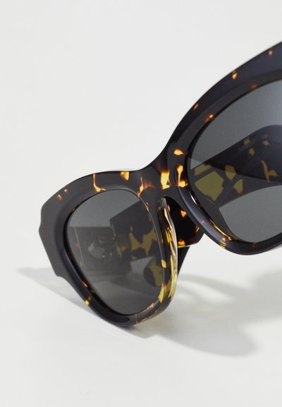 Occhiali da sole oversize con una fantasia tartarugata nera e marrone. Lenti grigie scure, montatura angolare e bracci spessi e ben delineati.