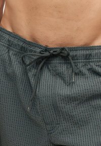 Torse masculin portant un short de bain texturé sombre noué par un cordon à la taille, montrant une partie du bas-ventre.