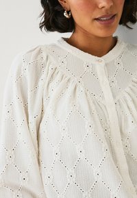 Camicia bianca con bottoni, caratterizzata da un tessuto testurizzato con motivi a diamante e accenti floreali. Maniche lunghe con dettaglio arricciato sulle spalle.