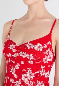 Rød blomstret tanktop med hvite blomstermønstre, teksturert stoff, formet halsutskjæring og tynne justerbare stropper.
