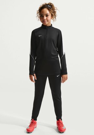 Młoda osoba ubrana w czarną bluzę treningową Nike i spodnie z białymi paskami oraz czerwone buty sportowe, stojąca i patrząca prosto na biały tle.