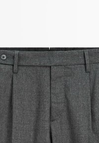 Pantalon gris foncé cintré avec passants de ceinture, plis et un seul bouton noir visible sur la ceinture.