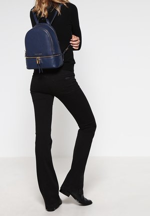 Marineblauwe leren rugzak met een afgeronde bovenkant, voorvak met rits en verstelbare schouderbanden, gedragen door een persoon in zwarte kleding en laarzen.