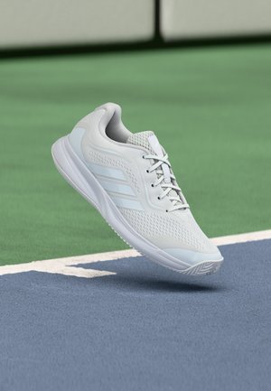 Baskets de sport blanches avec des rayures bleu clair flottant au-dessus d’un coin de terrain de sport extérieur bleu et vert.