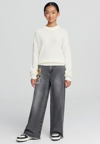 Maglione bianco a maglia con polsini a costine, abbinato a jeans grigi a gamba larga con una toppa a forma di orso marrone sulla parte anteriore, e sneakers in bianco e nero.