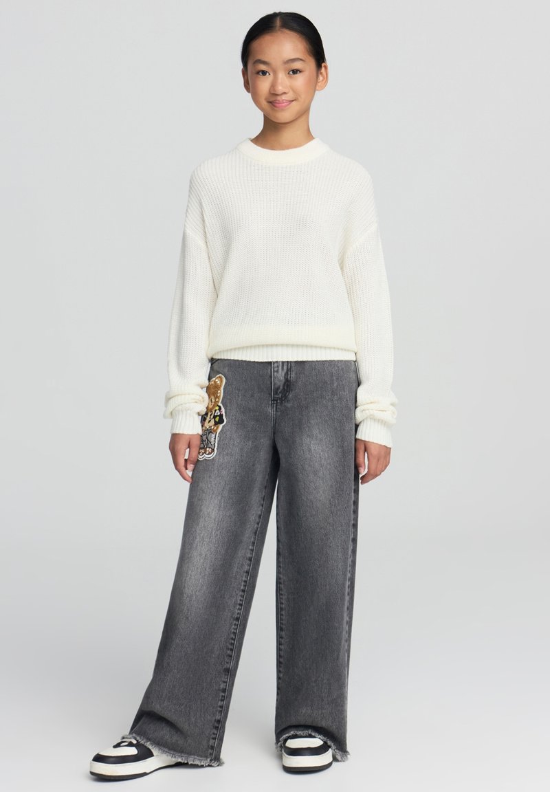 Maglione bianco a maglia con polsini a costine, abbinato a jeans grigi a gamba larga con una toppa a forma di orso marrone sulla parte anteriore, e sneakers in bianco e nero.