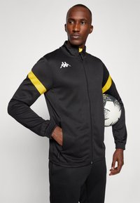 Black zip-up athletic jacket with yellow shoulder stripes, featuring a ribbed hem and cuffs. The fabric appears smooth and durable.