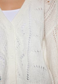 Bílý pletený cardigan s volným střihem, který má výstřih do V, zapínání na knoflíky a složité vzory. Měkká textura s otevřenými pletenými detaily.