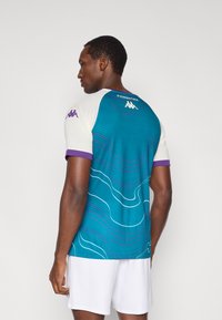 Majica s kratkimi rokavi v modrozeleni in beli barvi, z vijugastimi vijoličnimi črtami. Na rami je logotip Kappa, na hrbtu pa napis "FIORENTINA".