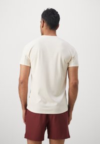Hombre vestido con una camiseta de manga corta en color blanco roto y pantalones cortos color granate, de pie de espaldas contra un fondo blanco liso.