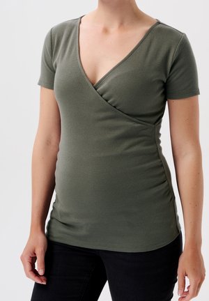 Oliemgroene top met korte mouwen, een V-hals en een wikkelontwerp. Geribbelde textuur met ruches aan de zijkant. Gecombineerd met zwarte broek.