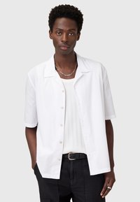 Camisa blanca de manga corta con cierre de botones en la parte delantera, fabricada con un tejido suave y de corte relajado, llevada sobre una camiseta sin mangas blanca texturizada.