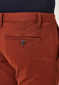 Rust-colored cotton chinos with a slim fit, featuring a back pocket with a black button, belt loops, and subtle logo stitching.