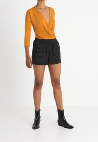 Hořčicově žlutý zavinovací top s třičtvrtinovými rukávy, v kombinaci s černými šortkami s bílými puntíky a černými kotníkovými botami.