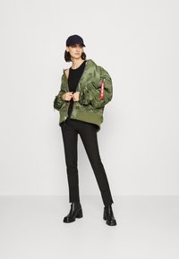 Green bomber jacket with zipper, orange lining, and red tag detail; worn over a black top and paired with tailored black pants and boots.