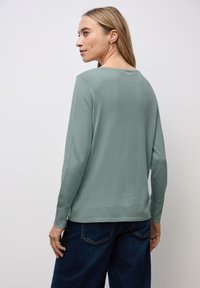 Langärmliges Oberteil aus gedämpftem grünen Stoff, mit Rundhalsausschnitt und gerippten Manschetten. Kombiniert mit dunklen Jeans. Rückansicht gezeigt.