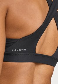 Svart sports-bh med et kryss-design på baksiden. Har logoen "CLOUDSPUN" i grått, laget av glatt, elastisk stoff.