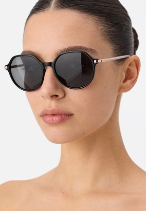 Vrouw met glad naar achteren gebonden haar, die zwarte ronde zonnebrillen met dunne metalen pootjes draagt, tegen een effen achtergrond.