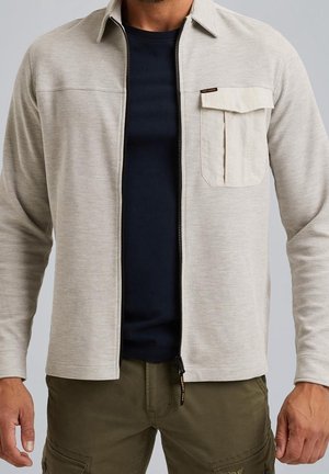 Mężczyzna w beżowej teksturowanej koszuli na zamek błyskawiczny, pod którą ma czarny t-shirt, i zielonych spodniach typu cargo, stoi na neutralnym tle.