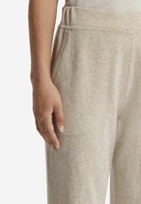 Ljust beige, mjukt tyg kvalitet joggingsbyxor med elastiskt midjeband, sidoficka och synliga sömdetaljer. Hand som vilar på byxorna.