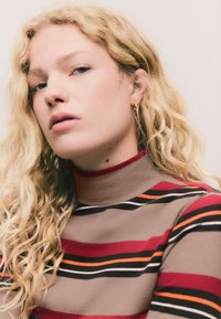 Garām piedurknēm ar augstu apkakli veidots džemperis bēša krāsā ar izteiksmīgām sarkanām, melnām un oranžām svītrām; mīksta tekstūra un piegulošs dizains; smalkas teksturētas detaļas.