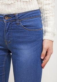 Vaqueros de denim azul con un diseño ajustado, que cuentan con un cierre frontal de botones, sutiles desgastes y detalles de costura en los bolsillos.