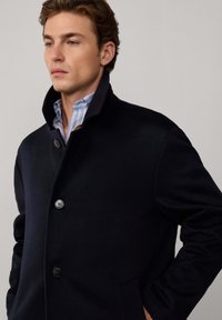 Giovane con capelli ricci che indossa un cappotto nero con bottoni sopra una camicia a righe blu, posando contro uno sfondo chiaro e uniforme.