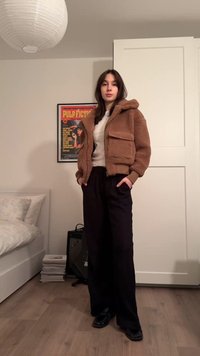 Bruine fleecejack met capuchon en zakken aan de voorkant, gecombineerd met zwarte broek met wijde pijpen en zwarte schoenen. Kamer met lichte houten vloer en beddengoed.