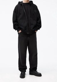 Personne portant un sweat à capuche noir ample avec fermeture éclair, un t-shirt noir, un jean noir ample et des chaussures noires à lacets, debout les mains dans les poches.