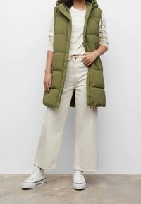 Oliwkowa pikowana kamizelka z kapturem, noszona na białej, w paski koszuli z długim rękawem. Do tego szerokie kremowe spodnie i białe trampki.