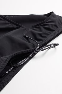 Débardeur de sport noir en tissu lisse et extensible. Il présente un col en V, des détails froncés et un cordon de serrage avec un texte imprimé.