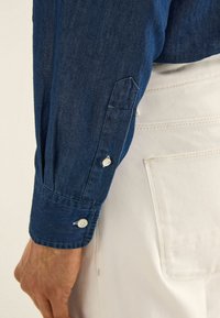 Camicia in denim a maniche lunghe, di colore blu scuro, con polsini abbottonati e una morbida texture, abbinata a pantaloni bianchi in tessuto con tasca.
