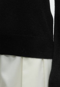 Ourlet de pull en maille noire sur jupe plissée blanche, montrant le contraste de textures entre les tissus.