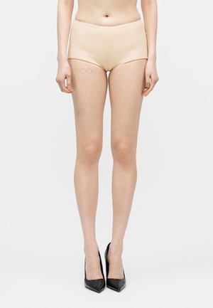 Žena nosiaca béžové vysoké šortky a čierne špicaté vysoké podpätky, s jemným tetovaním nekonečna na ľavom stehne.