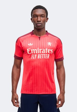 Jeune homme portant un maillot de football rouge Adidas avec « Emirates Fly Better » et un blason lion, debout devant un fond blanc.