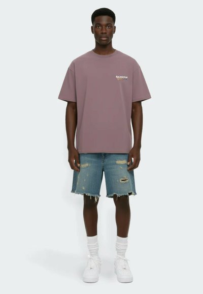 Young man stands wearing a mauve oversized T-shirt with small logo, distressed denim shorts, white socks, and white sneakers on plain background.