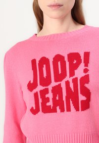 Roza pletena sv sweater s odvažnim crvenim grafičkim tekstom "JOOP! JEANS" istaknuto prikazanim na prednjem dijelu, s rebrastim izrezom i rubom.