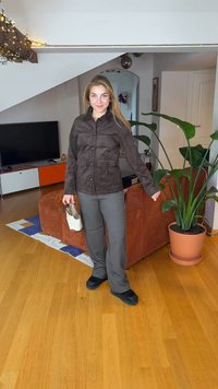 Καφέ τζάκετ από σουέτ με κουμπιά, γκρι παντελόνια με σχέδια, μαύρα παπούτσια και μια ανοιχτόχρωμη τσάντα. Το φόντο περιλαμβάνει ένα πράσινο φυτό και έναν καναπέ.