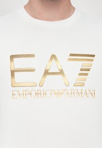Hvit bomull t-skjorte med metallisk gulllogo som leser «EA7 EMPORIO ARMANI», med rund hals og glatt tekstur.