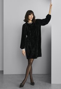 Rochie din catifea neagră cu mâneci lungi, țesătură texturată, croială lejeră și guler rotund. Purtați cu colanți negri și fundițe pe pantofi.