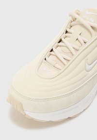 Nike Air sportbačiai šviesiai smėlinės spalvos, su tekstūruota medžiagos viršumi, apvaliu pirštu ir baltu tarpiniu padu. Subtilus logotipas šone ir ant liežuvėlio.
