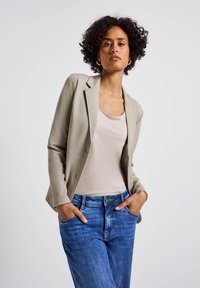 Blazer ajustado gris claro sobre una blusa beige, combinado con jeans de mezclilla azul. El blazer cuenta con un cuello con muesca y dos botones.