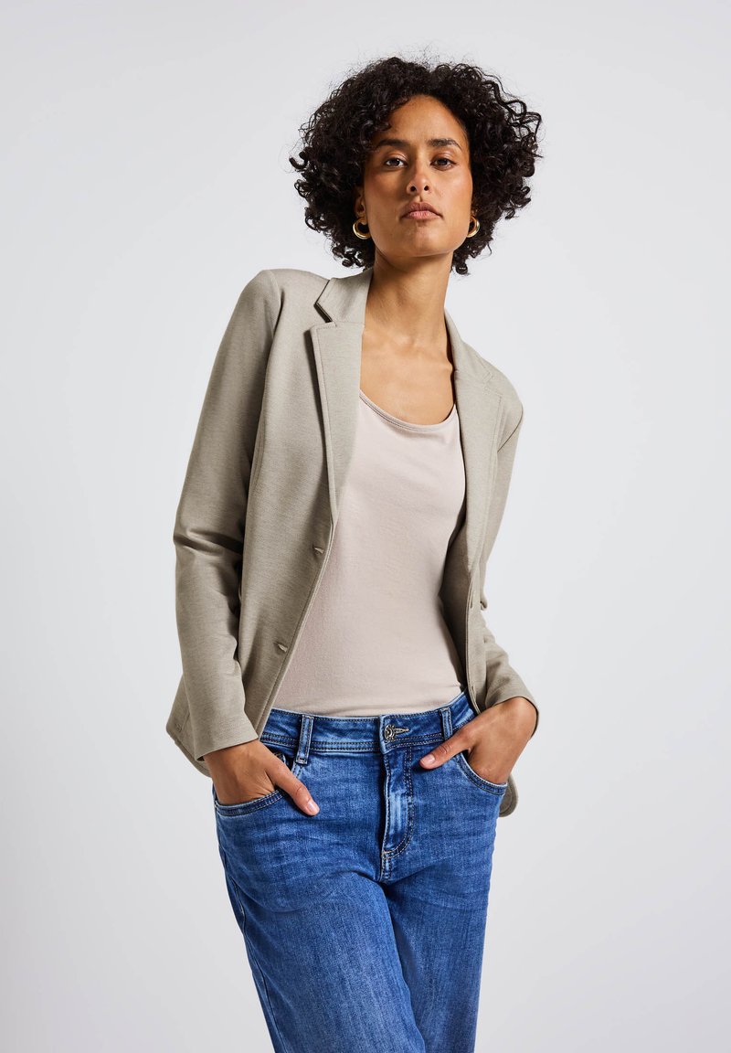 Blazer ajustado gris claro sobre una blusa beige, combinado con jeans de mezclilla azul. El blazer cuenta con un cuello con muesca y dos botones.