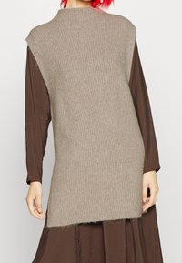 Gilet in maglia beige con scollo alto e ampie aperture per le braccia, caratterizzato da una texture a coste e un orlo posteriore più lungo. Indossato sopra un vestito marrone.