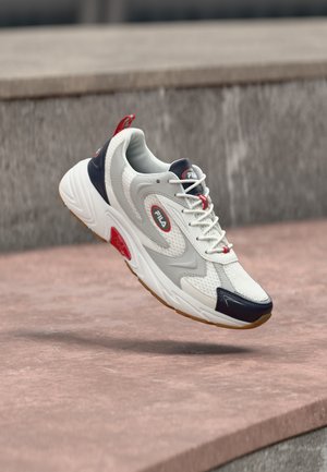 White, navy, and red FILA sneaker with mesh texture and rubber sole, suspended mid-air above a stone surface and steps.